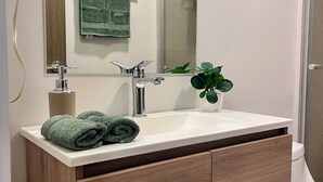 Ein modernes Badezimmer mit einem Holzbadezimmerhocker, einem weißen Waschbecken, einem Spiegel und einer grünen Pflanze.