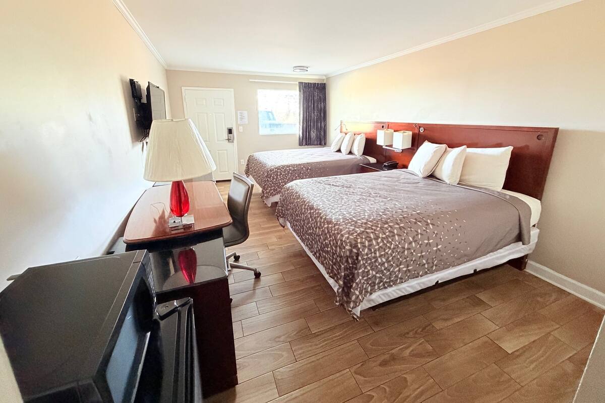 A hotel room with two beds, a desk with a lamp, a TV, and a wooden floor.