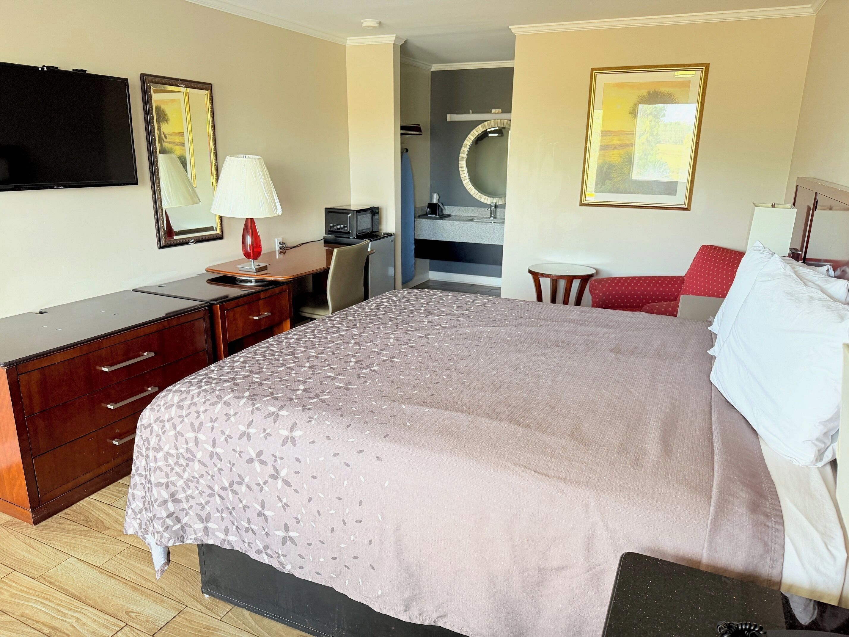 Habitación de hotel con cama, escritorio, silla, televisor, lámpara, espejo y baño visible a través de una puerta abierta.