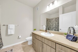 Un baño con encimera color beige, lavamanos blanco y un espejo grande. Hay un perchero con una toalla blanca y una pequeña maceta sobre la encimera.