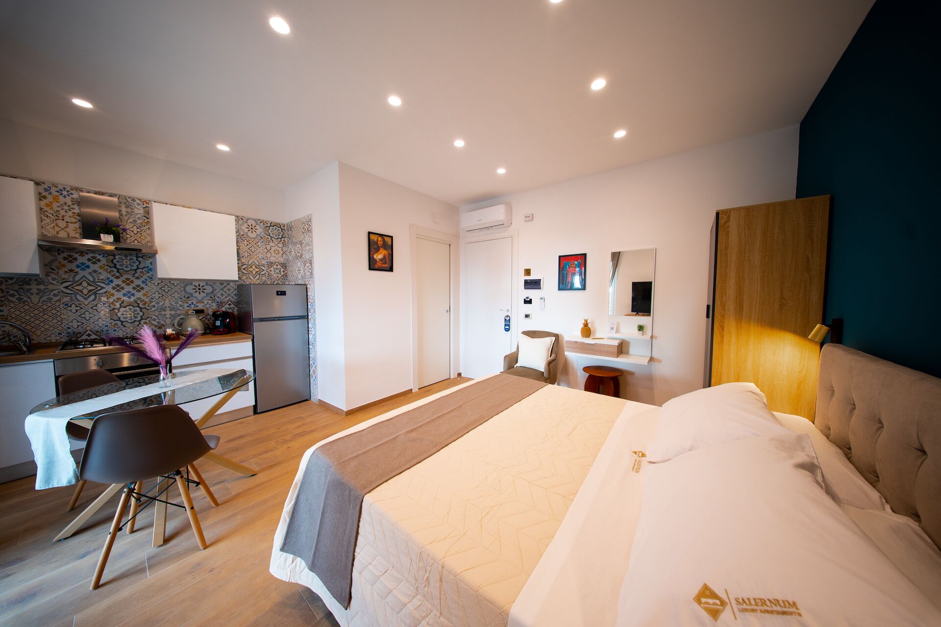 A modern hotel room with a bed, a desk, a kitchenette, and a bathroom.