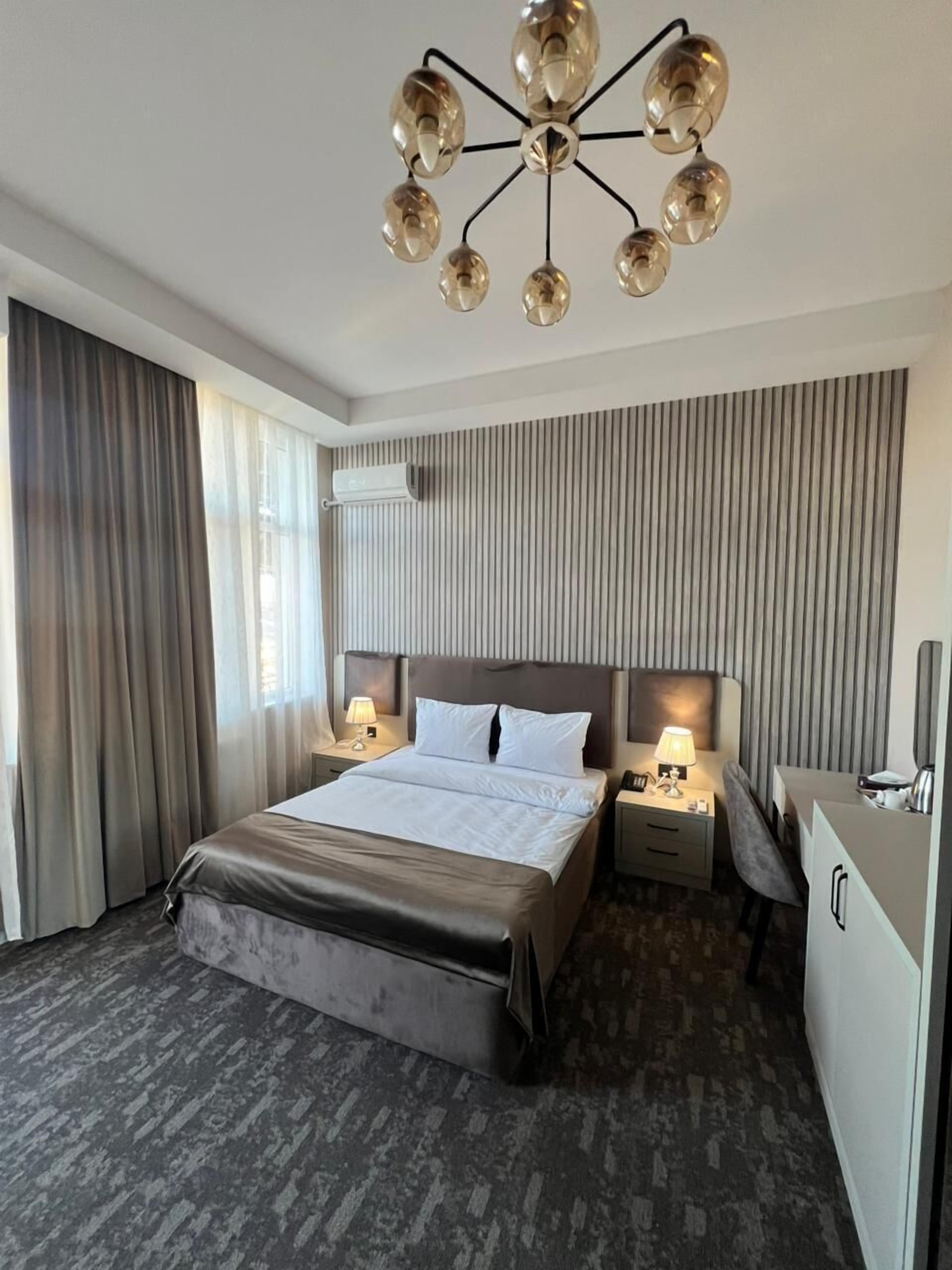 A hotel room with a large bed, two bedside tables with lamps, a desk with a chair, a wardrobe, and a modern chandelier.
