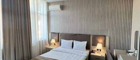A hotel room with a large bed, two bedside tables with lamps, a desk with a chair, a wardrobe, and a modern chandelier.