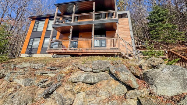 Une cabane moderne à deux étages, avec une façade en bois, un balcon à l’étage supérieur et des fondations en pierre.