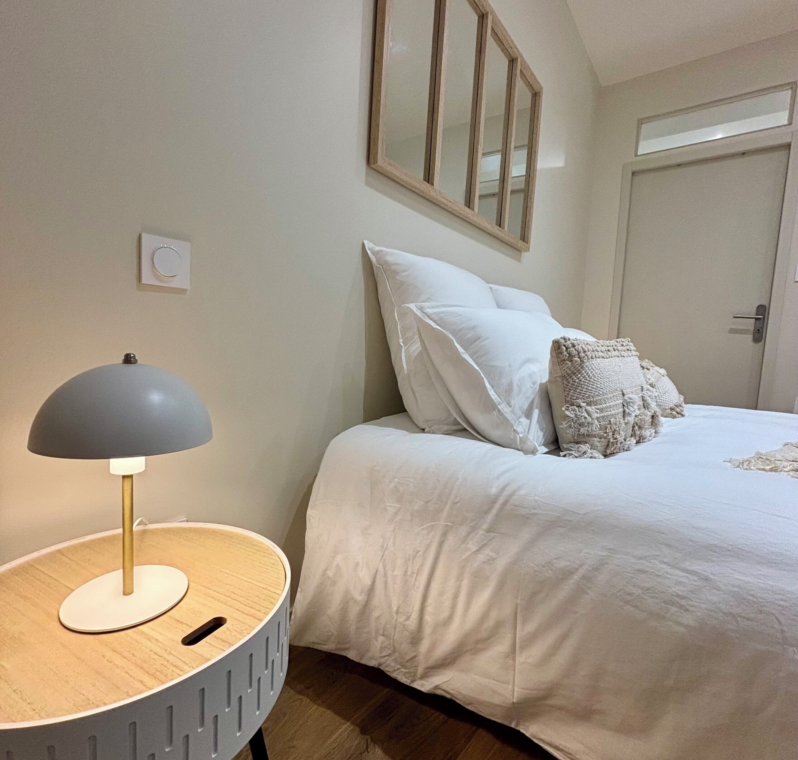 A bedroom with a bed, a round side table with a lamp, a mirror on the wall, and a door.