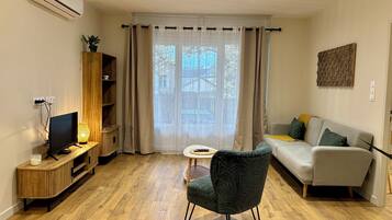 Ett modernt vardagsrum med en platt-TV, ett litet runt bord, en grön klädd fåtölj och en soffa med kuddar.
