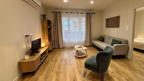 A modern living room with a wooden floor, a sofa, a chair, a TV, and a small table. - Casa Koenig - T2 spacieux & Terrasse en hyper-centre (Brive-la-Gaillarde)