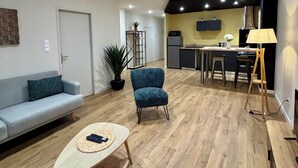 A modern living room with a sofa, a coffee table, a blue armchair, and a kitchen area with a dining table, chairs, and a lamp. - Casa Koenig - T2 spacieux & Terrasse en hyper-centre (Brive-la-Gaillarde)