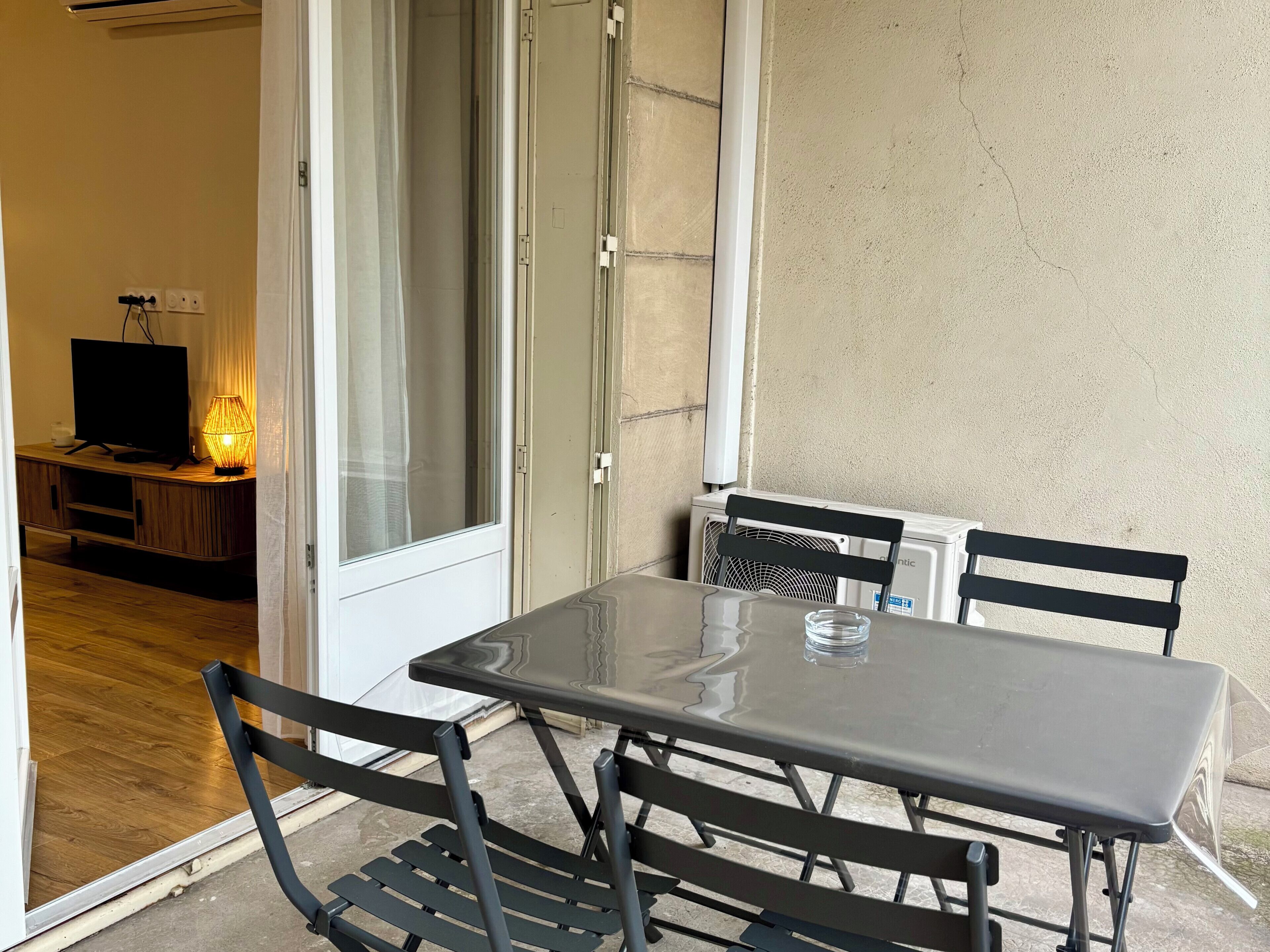 Una habitación con puerta de vidrio que da a un balcón, una mesa con sillas, un televisor sobre un mueble de madera y un aire acondicionado.