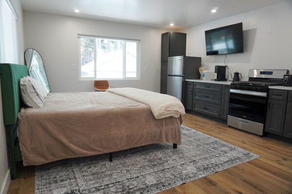 Quarto com cama, cadeira, janela, televisão de tela plana e cozinha equipada com geladeira, fogão e micro-ondas.
