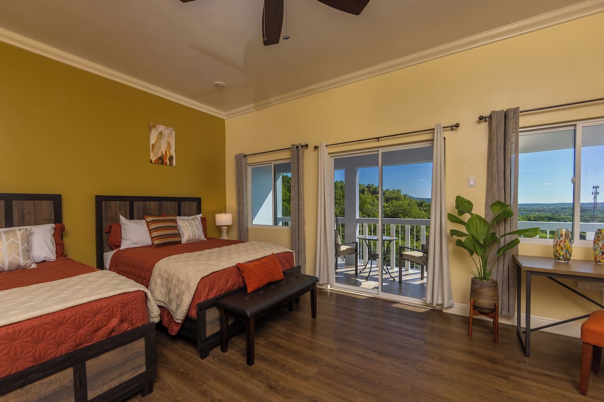 Habitación de hotel con dos camas, un banco, una mesa y un balcón con vistas.