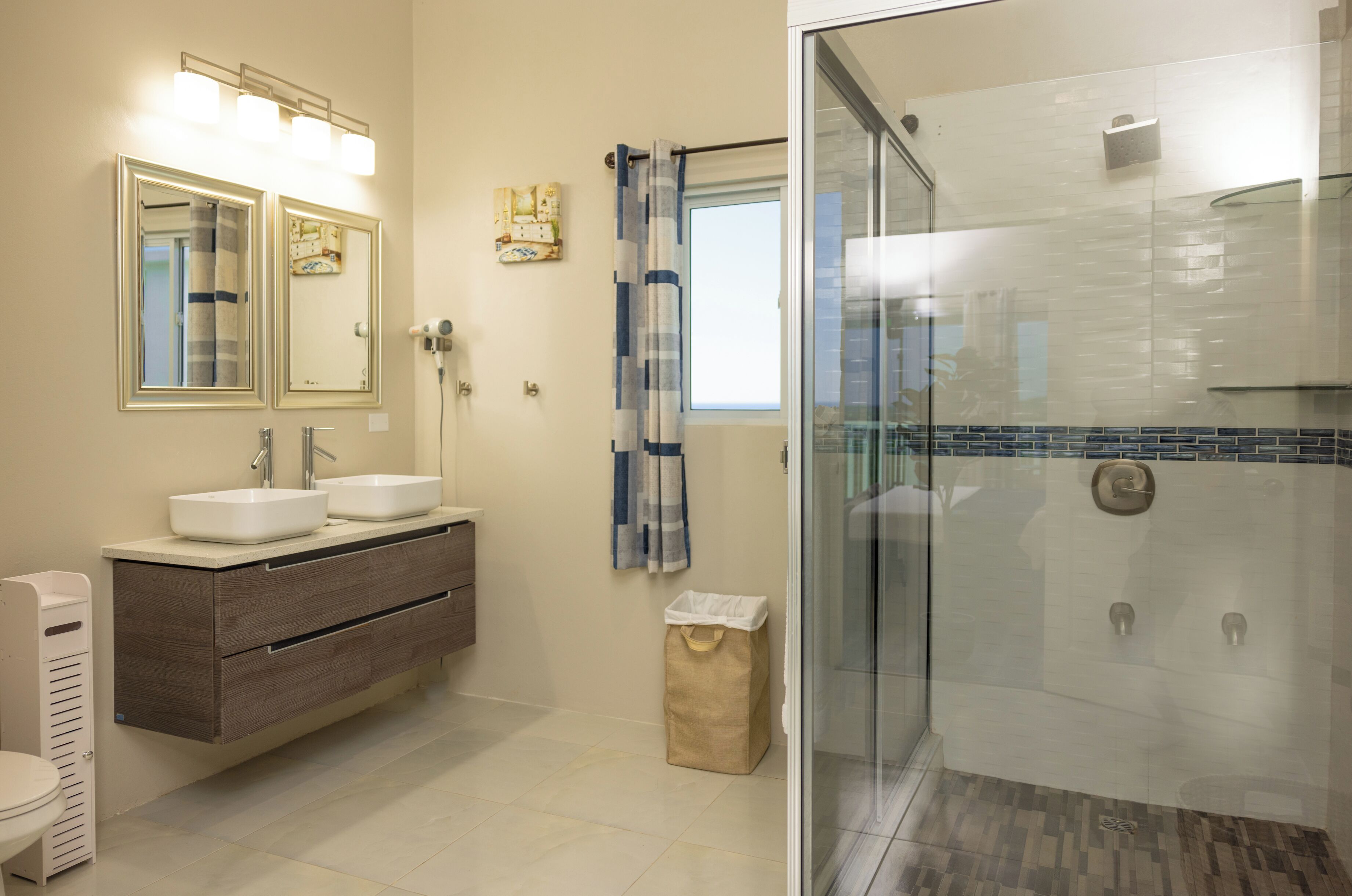Un baño con dos lavamanos, un tocador, ducha con puerta de vidrio y un pequeño cubo de basura.
