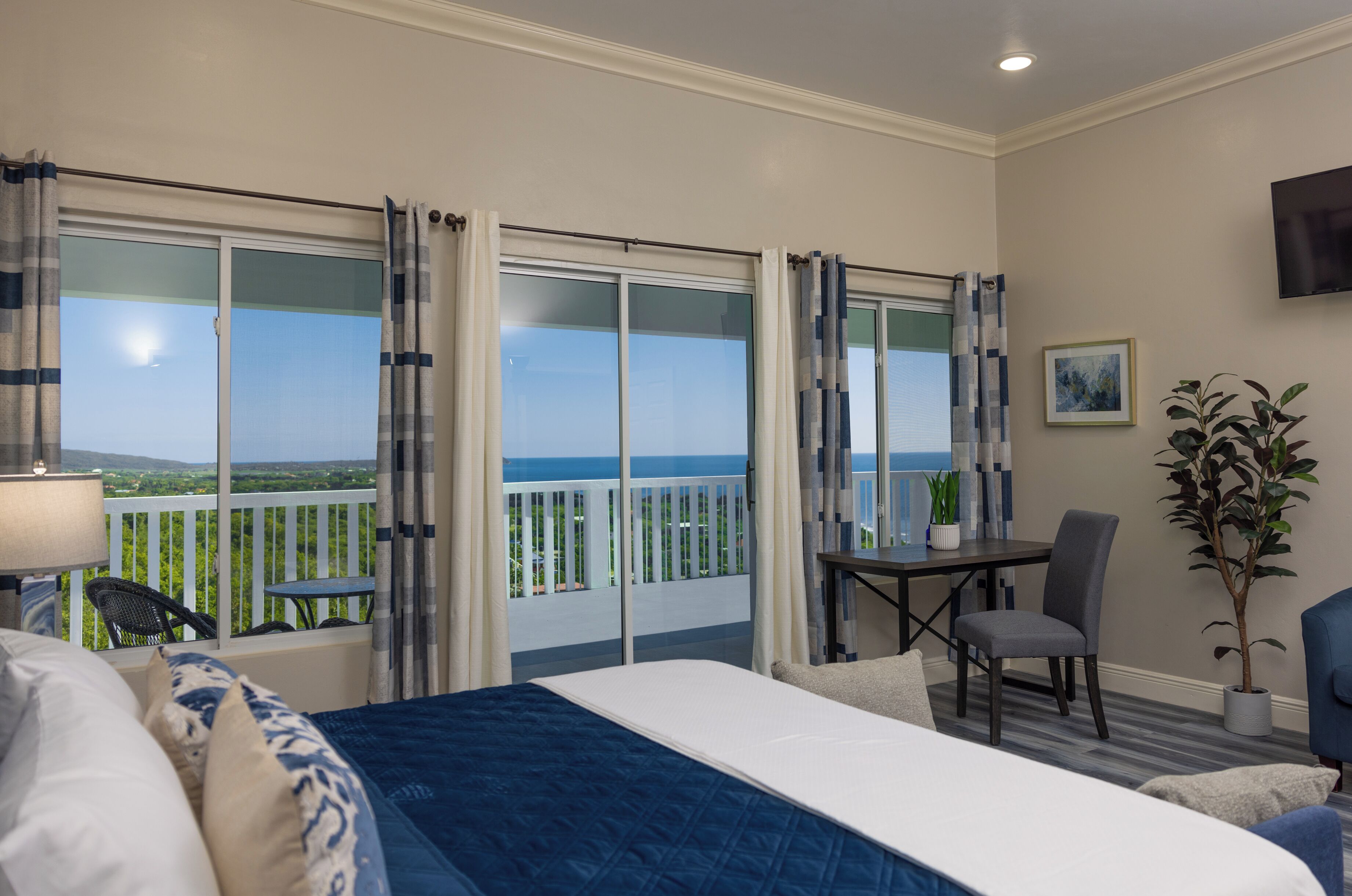 A hotel room with a bed, a desk, a chair, a potted plant, and a balcony with a view of the ocean.