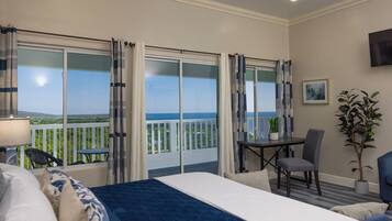 A hotel room with a bed, a desk, a chair, a potted plant, and a balcony with a view of the ocean.