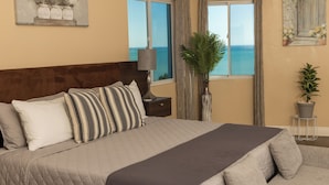 Un dormitorio con una cama grande, una mesita de noche, una lámpara, una planta en maceta y vista al mar a través de una ventana.