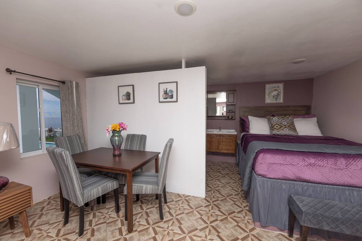 Una habitación de hotel compacta con comedor, una cama con colcha morada y una pequeña kitchenette.