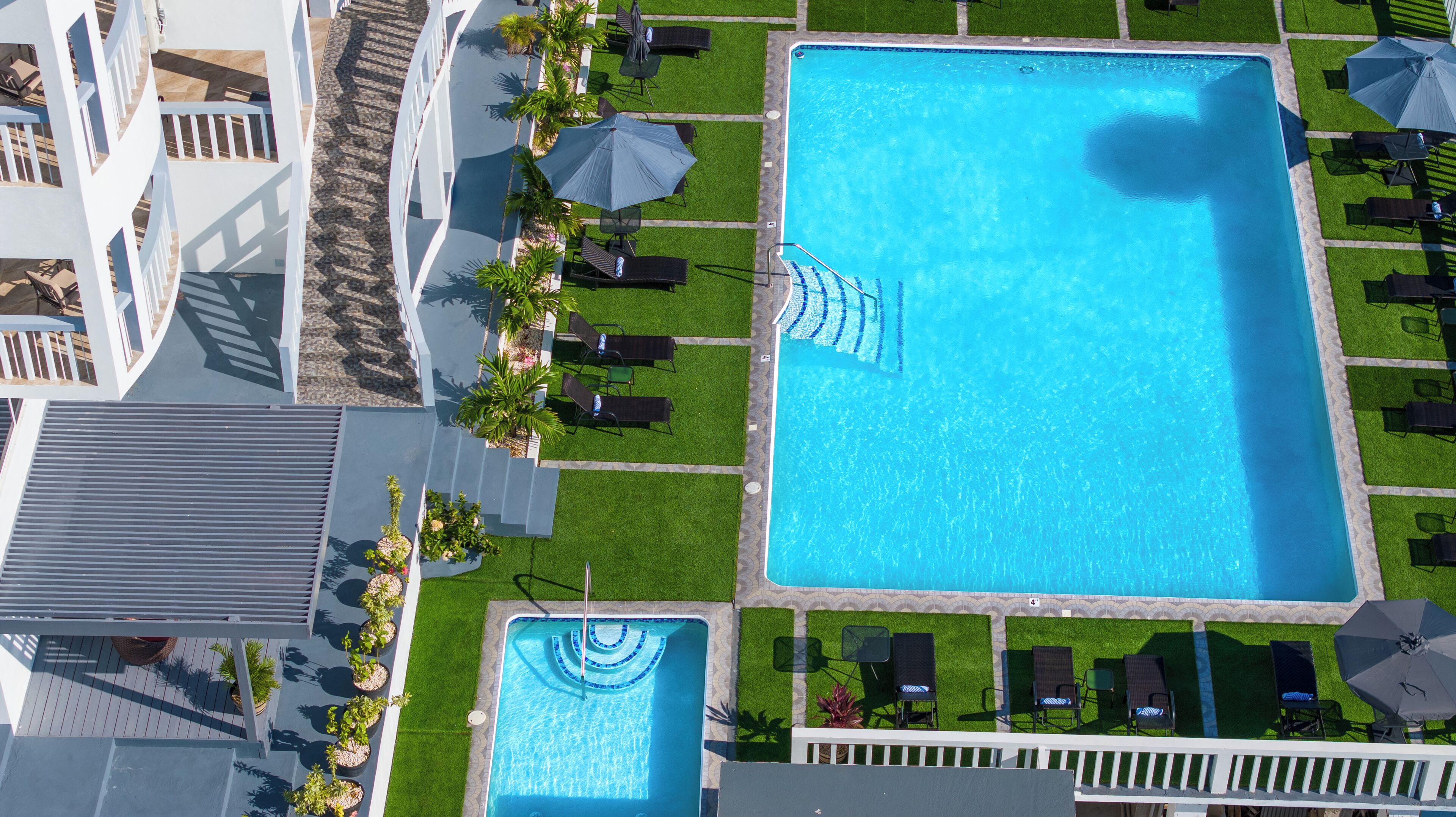 Una gran piscina rectangular con una piscina más pequeña contigua, rodeada de sillas de jardín y sombrillas, con una cerca blanca y un edificio al fondo.