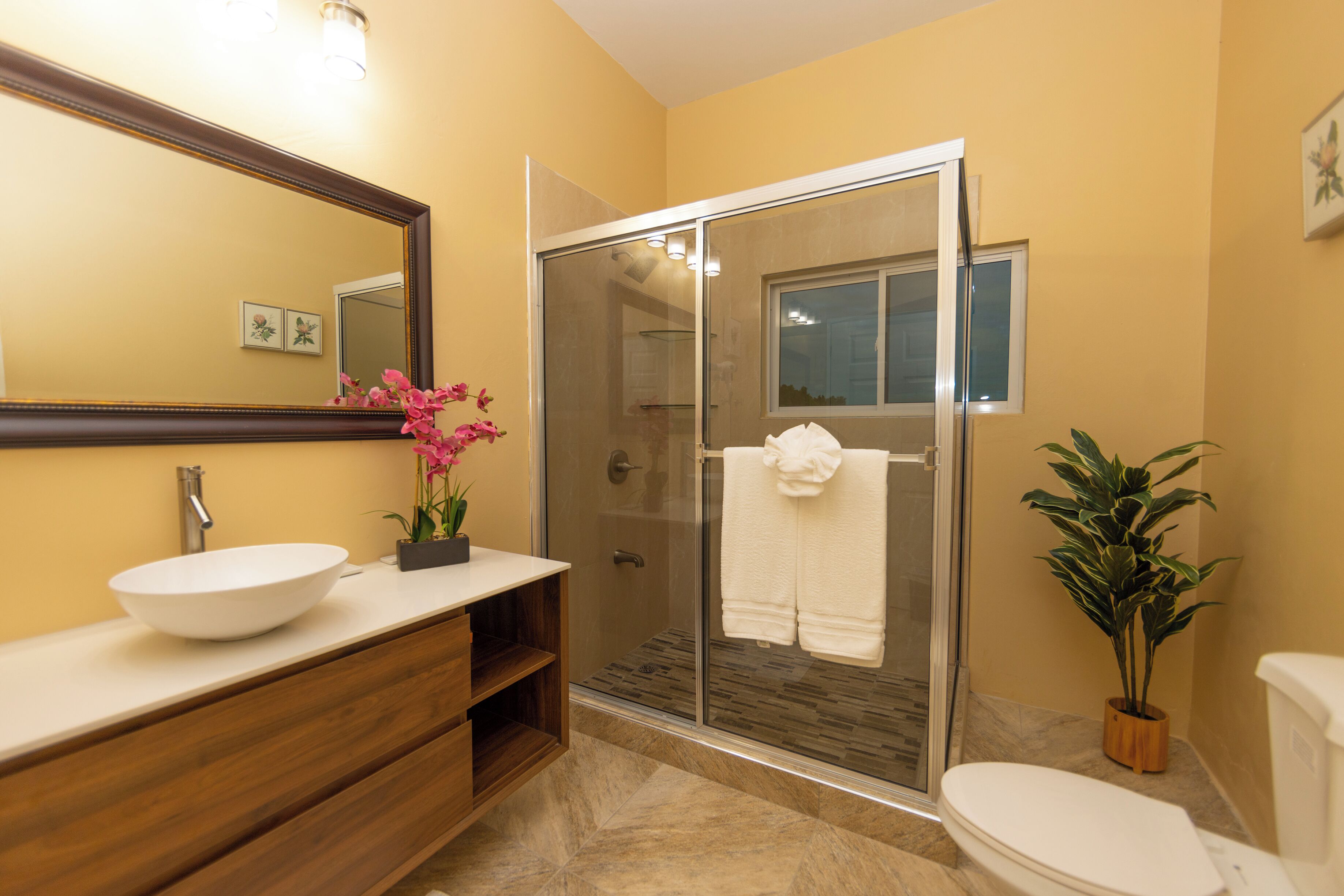 A bathroom with a white sink, wooden vanity, a glass shower enclosure, and a potted plant.