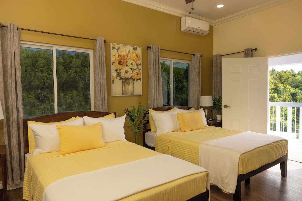Habitación de hotel con dos camas, un ventanal con cortinas, un cuadro de flores y una puerta que da a un balcón.