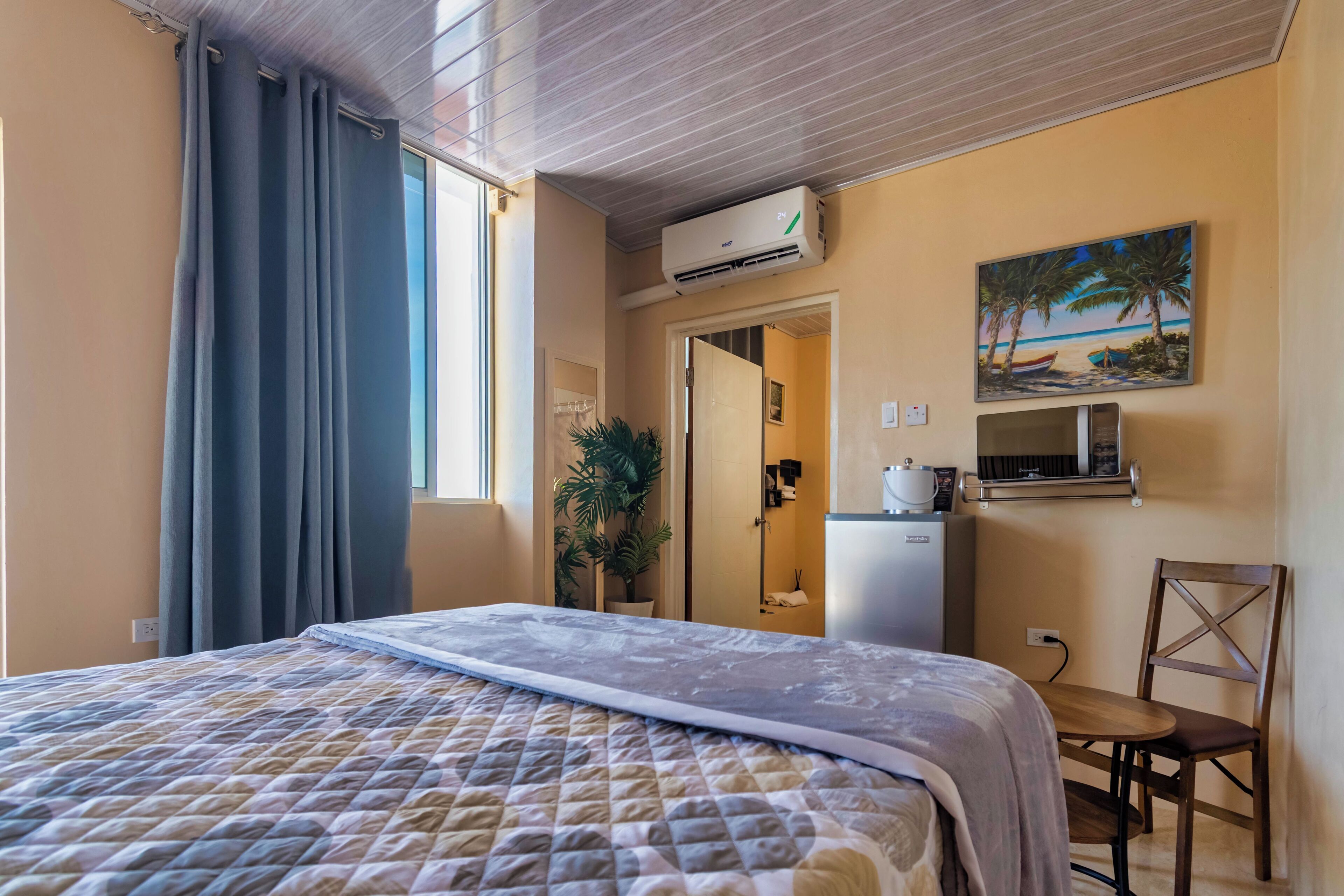A hotel room with a bed, a desk, a chair, an air conditioning unit, a painting, and a potted plant.