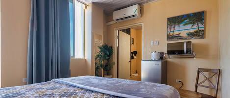 A hotel room with a bed, a desk, a chair, an air conditioning unit, a painting, and a potted plant.