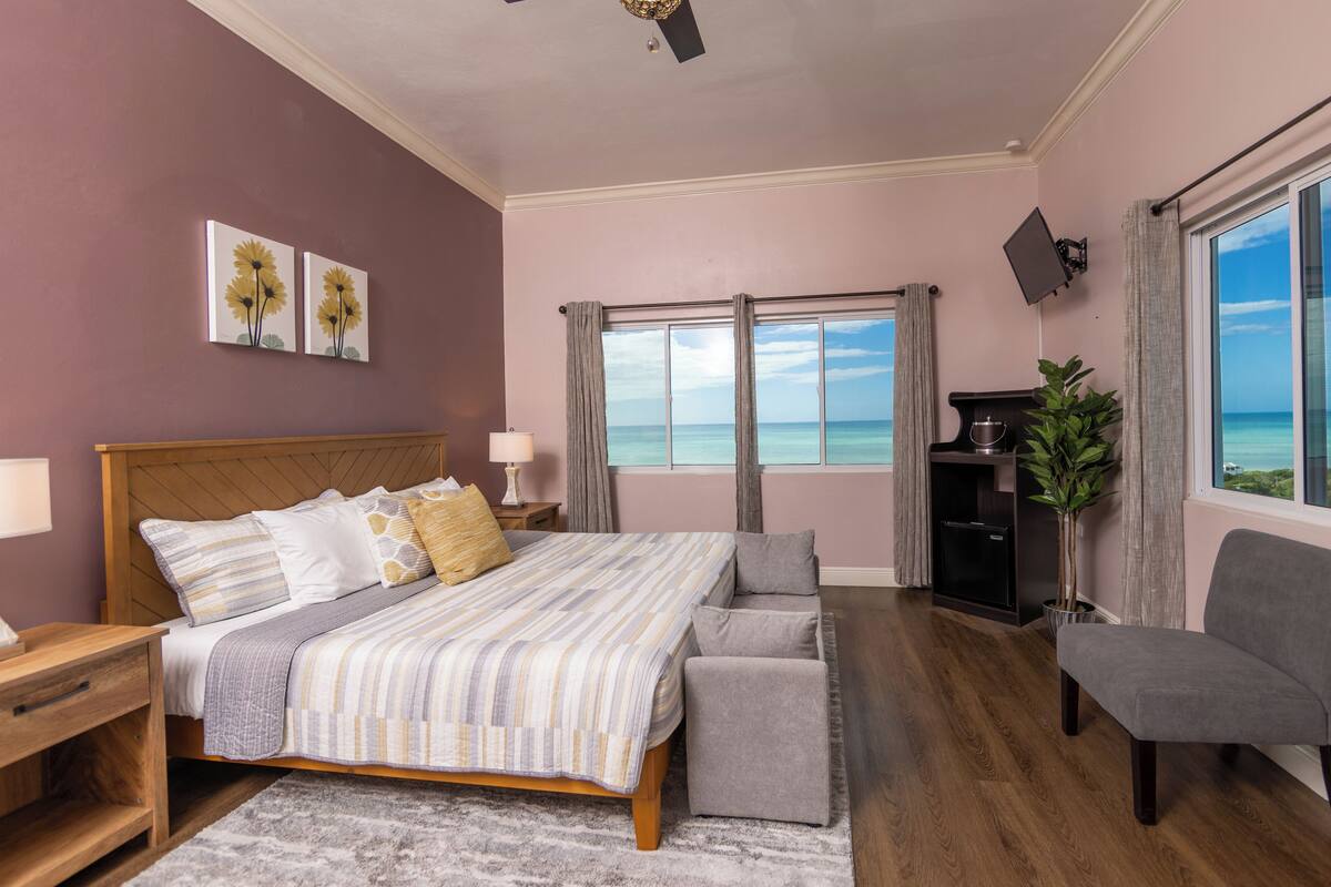 Un dormitorio con cama, mesitas de noche, una silla, escritorio, planta y vista al océano.