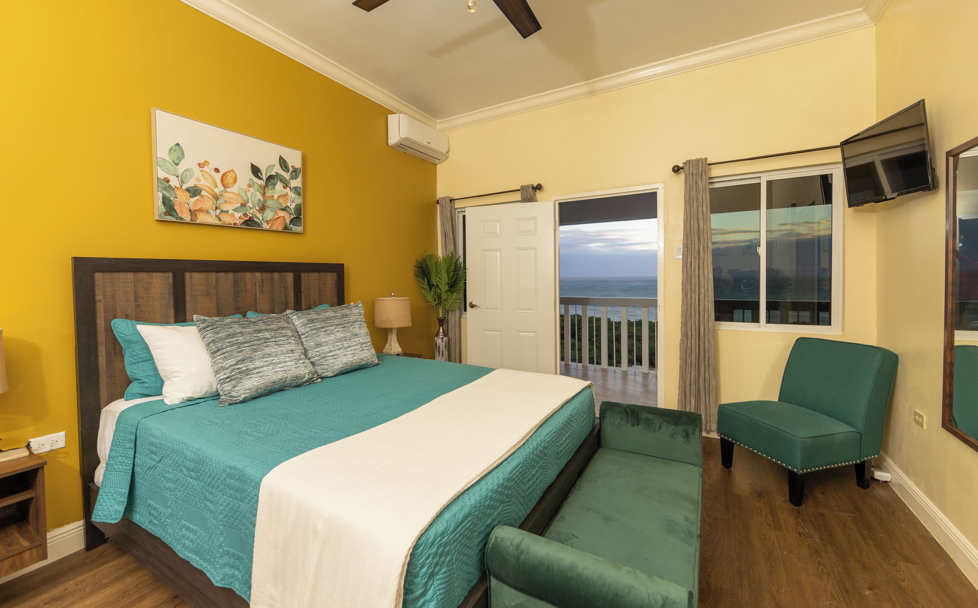 A bedroom with a bed, a chair, a TV, and a door leading to a balcony with a view of the ocean.
