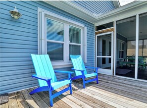 A covered porch with two blue Adirondack chairs, a sliding glass door, and a window with blinds. - "Hooked For Life" Cozy 3 Bedroom/2 Bath Getaway w/ Hot Tub. 1 Min. Walk to Beach (Oak Island)