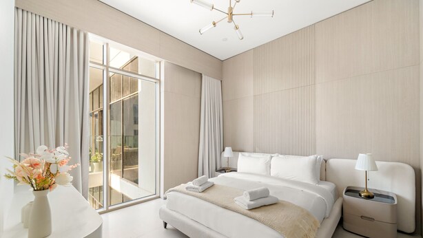 Quarto de hotel moderno com uma cama grande, mesa de cabeceira com abajur, um vaso de flores e vista para o exterior por uma porta deslizante.