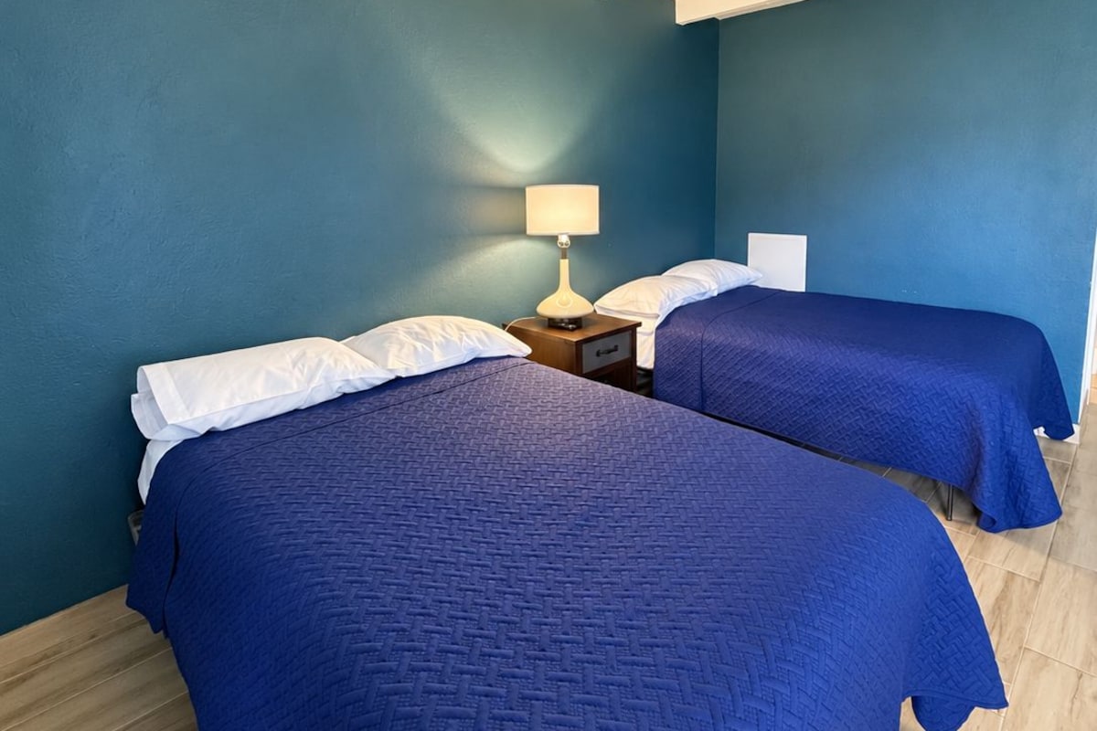 A room with two beds, a nightstand with a lamp, and a blue wall.