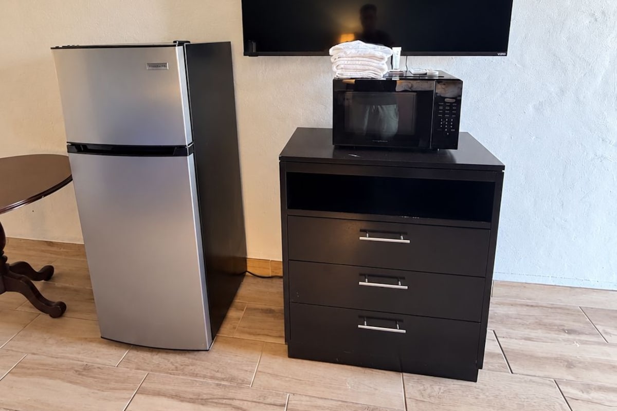 A kitchenette with a stainless steel refrigerator, a microwave on a black cabinet, and a flat-screen TV mounted on the wall.