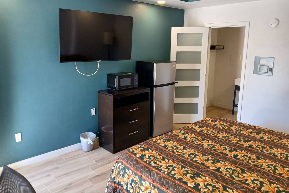 A bedroom with teal walls, a bed with a patterned bedspread, a black TV mounted on the wall, a white door, and a small cabinet with a microwave on top.
