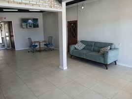 Uma sala ampla com um sofá cinzento, duas cadeiras de escritório, uma mesa de madeira e uma TV fixada na parede.