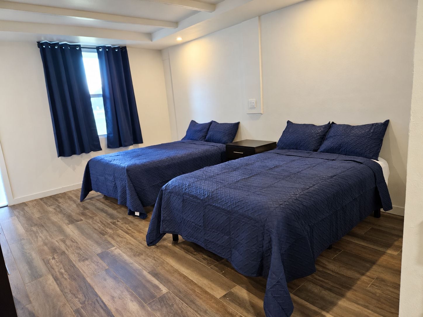 Una habitación de hotel con dos camas, una ventana con cortinas, una mesita de noche y piso de madera.