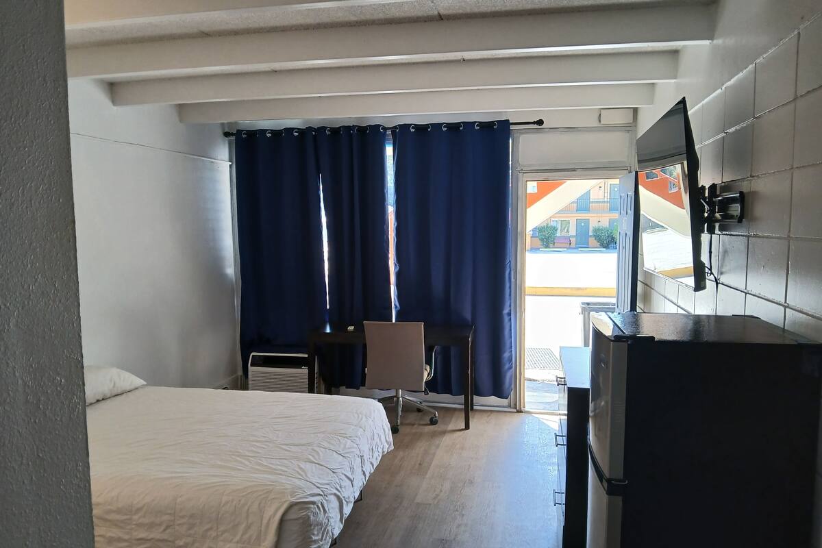 A hotel room with a bed, a desk, a chair, a TV, and a large window with curtains.