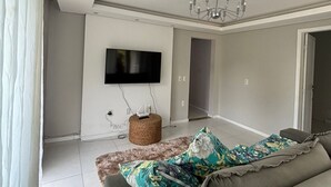 A modern living room with a grey sofa, a flat-screen TV mounted on the wall, a chandelier, and a wicker ottoman. - Beautiful penthouse with a view. (Florianópolis)