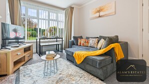 Ein modernes Wohnzimmer mit grauem Sofa, einem HolztV-Schrank, einem kleinen runden Tisch und einem großen Fenster mit Vorhängen.