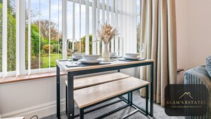 Ein modernes Zimmer mit einem großen Fenster, einem Esstisch gedeckt mit weißen Tellern und Schüsseln, einer Bank und einer Vase mit getrockneten Pflanzen.
