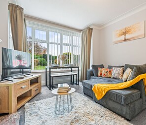 A modern living room with a grey sofa, a wooden TV stand, a small round table, and a large window with curtains.