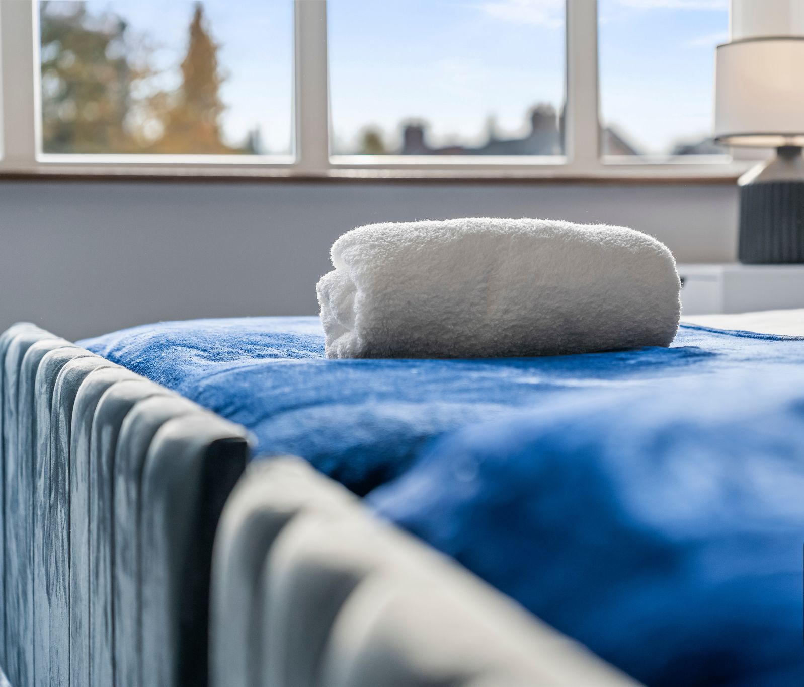 Uma cama bem arrumada com um cobertor azul e uma toalha branca, um abajur e uma janela com vista para árvores e edifícios.