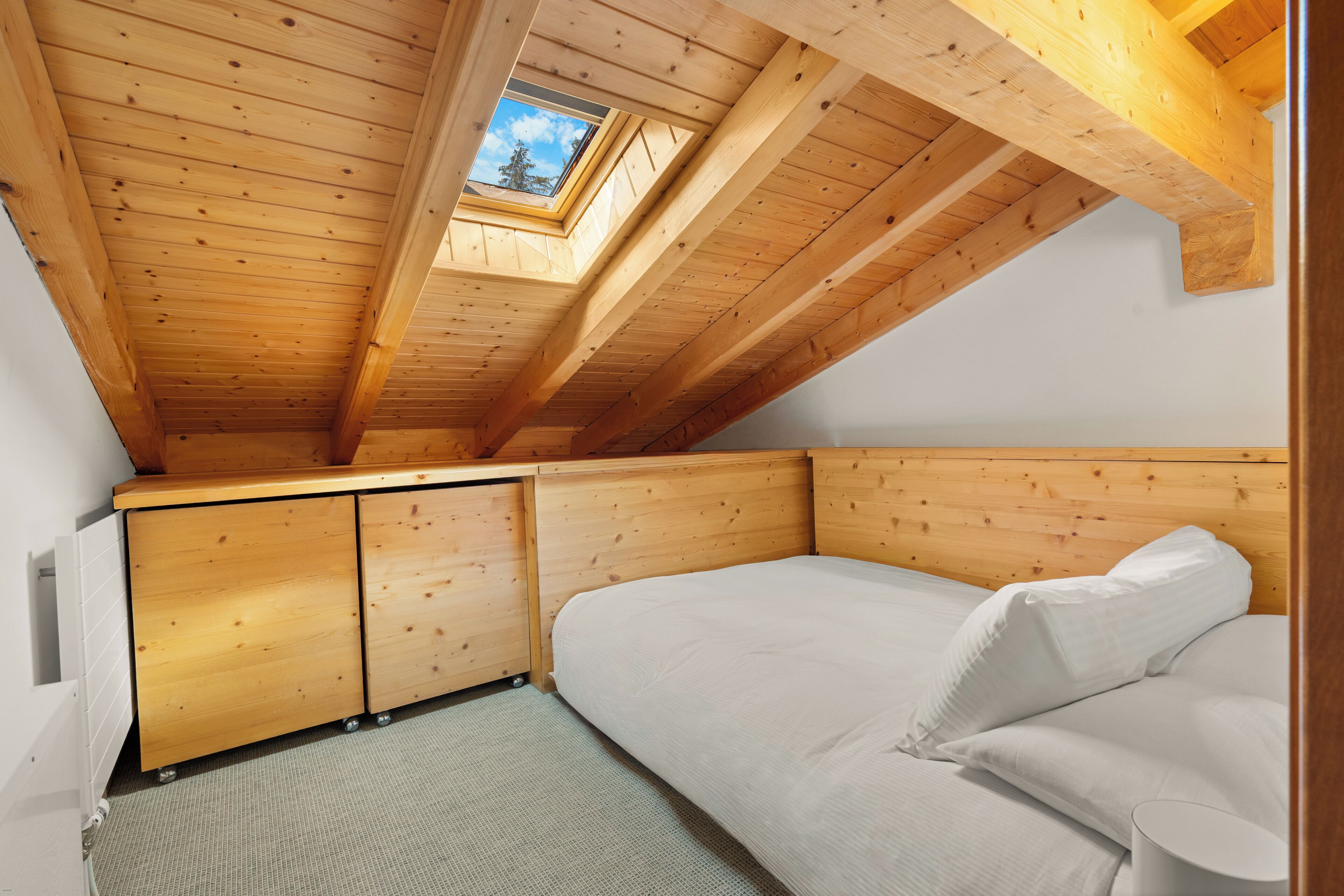 A room with a sloped ceiling, wooden beams, a skylight, a bed, and a small cabinet.
