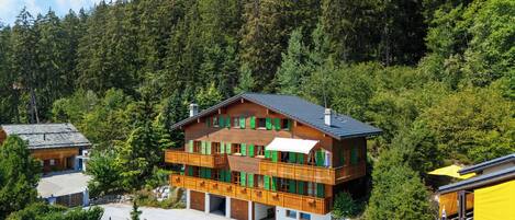 A multi-story wooden chalet with a balcony, set in a forested mountainous area.
