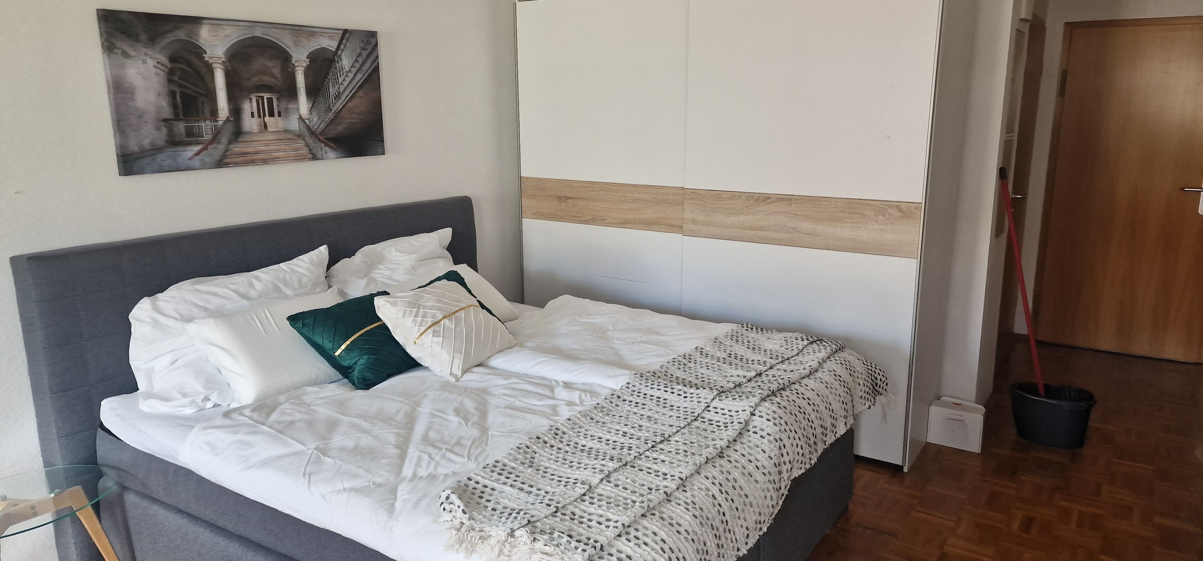 Een netjes opgemaakt bed met witte lakens, een grijze hoofdbord en een decoratieve gebreide plaid.
