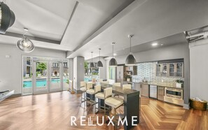 A modern kitchen with a central island, a dining area with bar stools, and a pool visible through glass doors. - Reluxme Luxury 2BR | Walk to Stadiums & Convention Center (Charlotte)