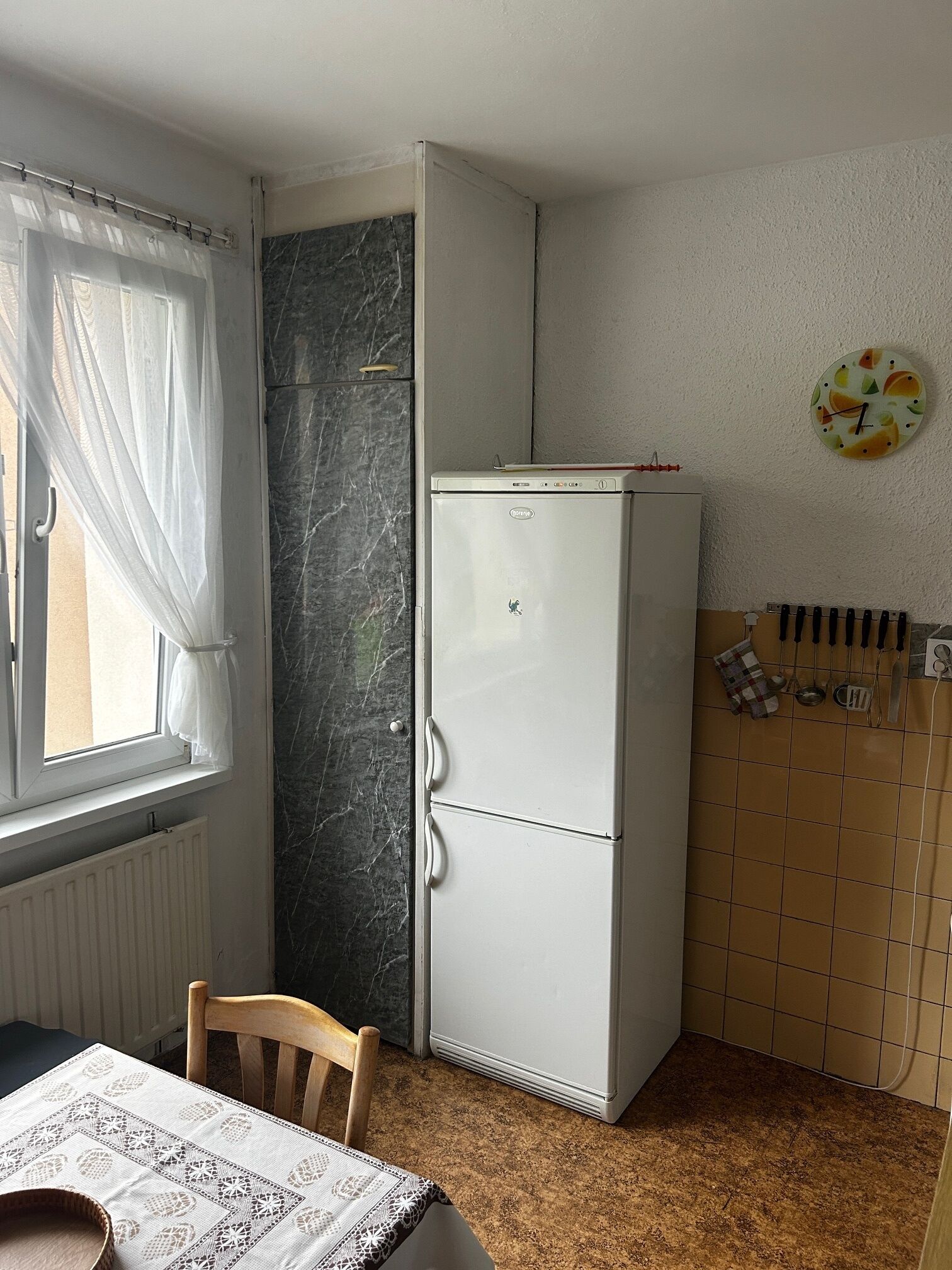 A compact kitchen with a refrigerator, a window with curtains, a tiled backsplash, and a wall-mounted clock.