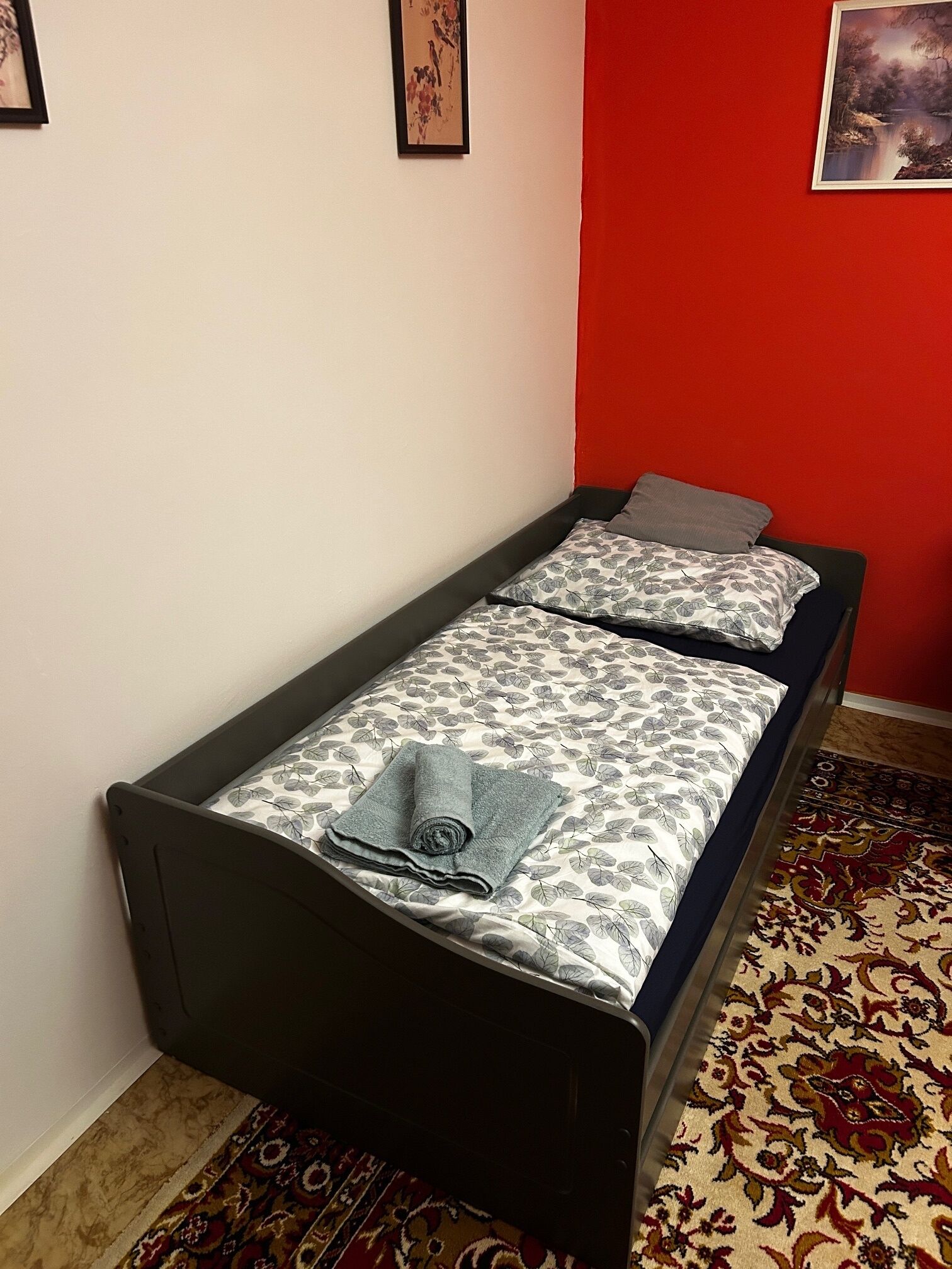 A single bed with a patterned bedspread and pillows, a rolled towel, and a red accent wall.
