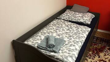 A single bed with a patterned bedspread and pillows, a rolled towel, and a red accent wall.