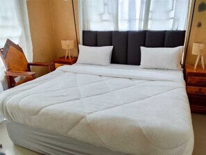 Una cama bien hecha con un edredón blanco acolchado, un cabecero tapizado oscuro y mesitas de noche de madera con lámparas.