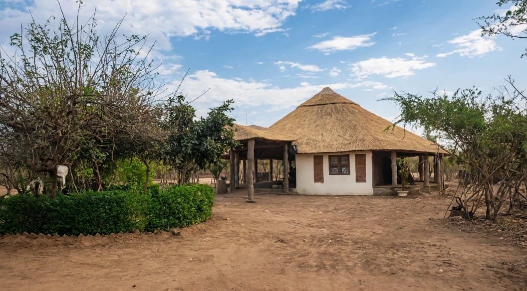 A rustic building with a thatched roof, surrounded by greenery and a dirt path.