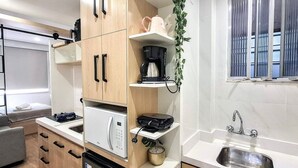 Cocina compacta con microondas, cafetera y lavabo. Hay una ventana con persianas y un teléfono en la pared.